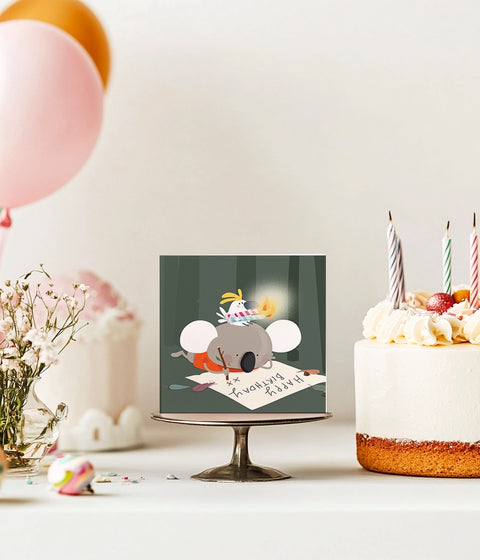 Square birthday card featuring a kawaii koala writing a to do list with a cockatoo and candles